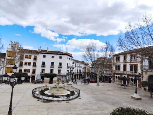 Plaza con fuente, edificios históricos y árboles bajo un cielo nublado en un pueblo español.